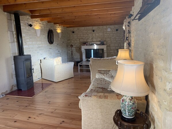 A cozy living room with a wood-burning stove, a sofa, a patterned armchair, and a lamp on a wooden stand. - La Grange – Spacious 3-Bedroom 19th-Century Barn with Private Garden (Le Coudreau Saint-Sigismond)