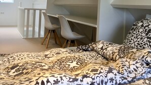 A modern bedroom with a sloped ceiling, a bed with patterned bedding, a desk with two chairs, and a window with a view of a brick building. - B&B depolderschuur is an independent home (Tienhoven)