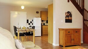 Uma sala de estar com sofá, uma mesa de jantar com cadeiras, uma cozinha com geladeira e armários, e uma escada.