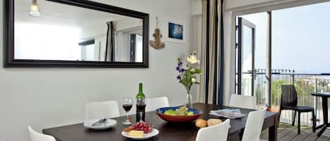 A dining area with a dark wooden table, white chairs, a mirror, a vase with flowers, and a view of the outdoors.