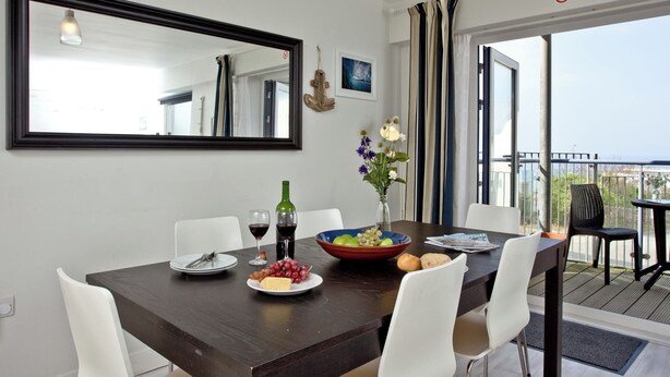 A dining area with a dark wooden table, white chairs, a mirror, a vase with flowers, and a view of the outdoors. - Apartment 7, Ocean 1 (Newquay)