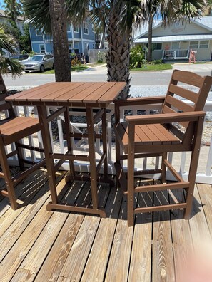 A wooden deck with a table and chairs, surrounded by a white fence, palm trees, and a building in the background.