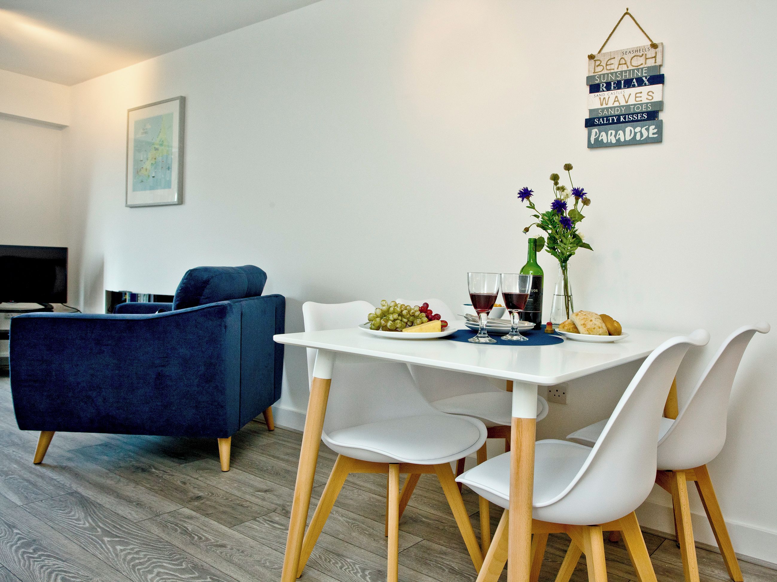 En matplats med ett vitt bord, två vita stolar, en blå soffa och en väggdekoration med ord som ”STRAND” och ”PARADIS”.