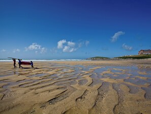 A beach with wet sand patterns, a building in the background, and a person carrying a surfboard.