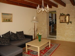 Ein Wohnzimmer mit Sofa, Couchtisch, einer Kandelaber und einem Gemälde, im Hintergrund eine Küche.