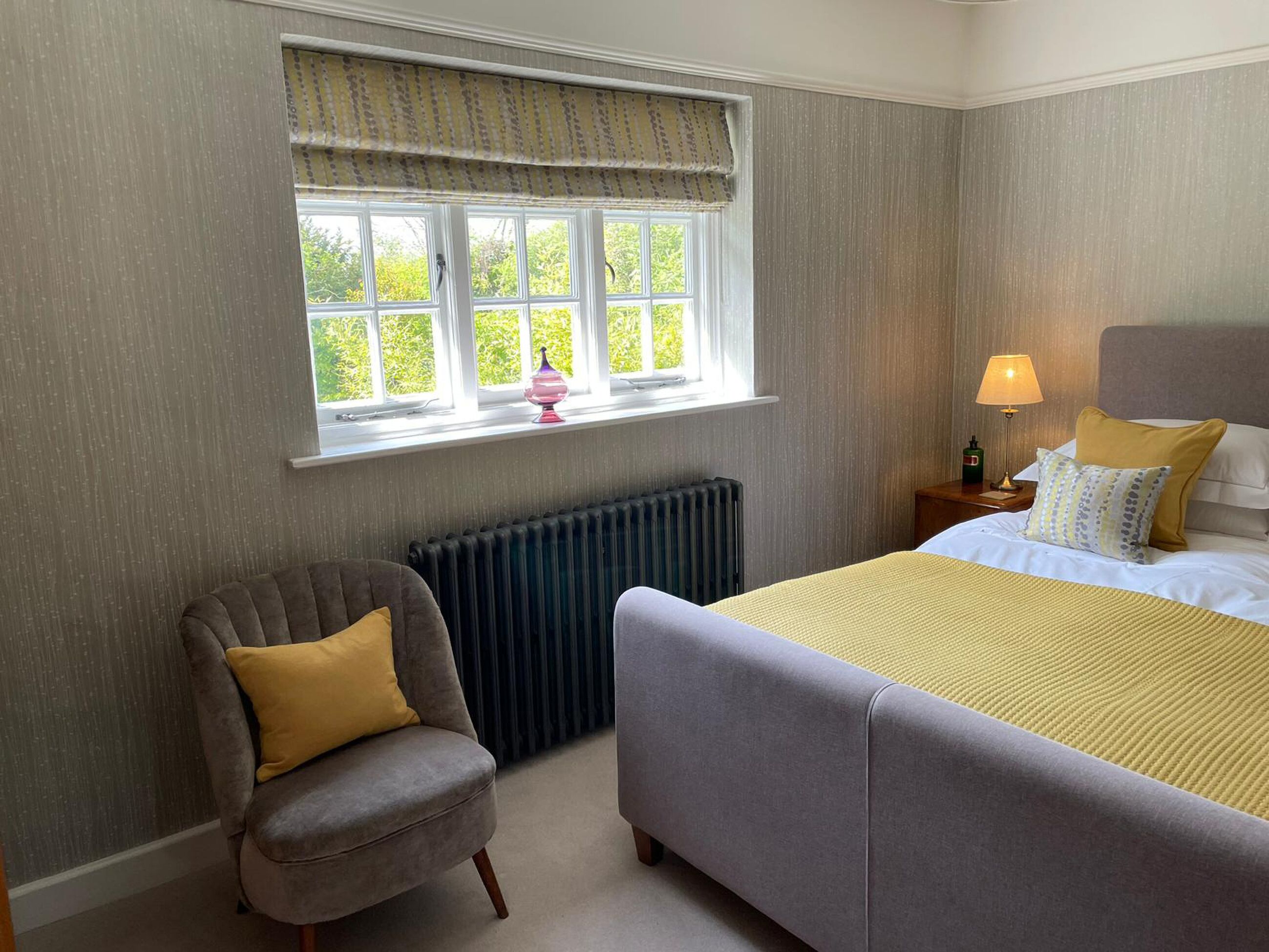 Ein Schlafzimmer mit einem Bett, einem Sessel, einem Fenster mit Vorhängen, einer Heizung und einem Nachttisch mit Lampe.