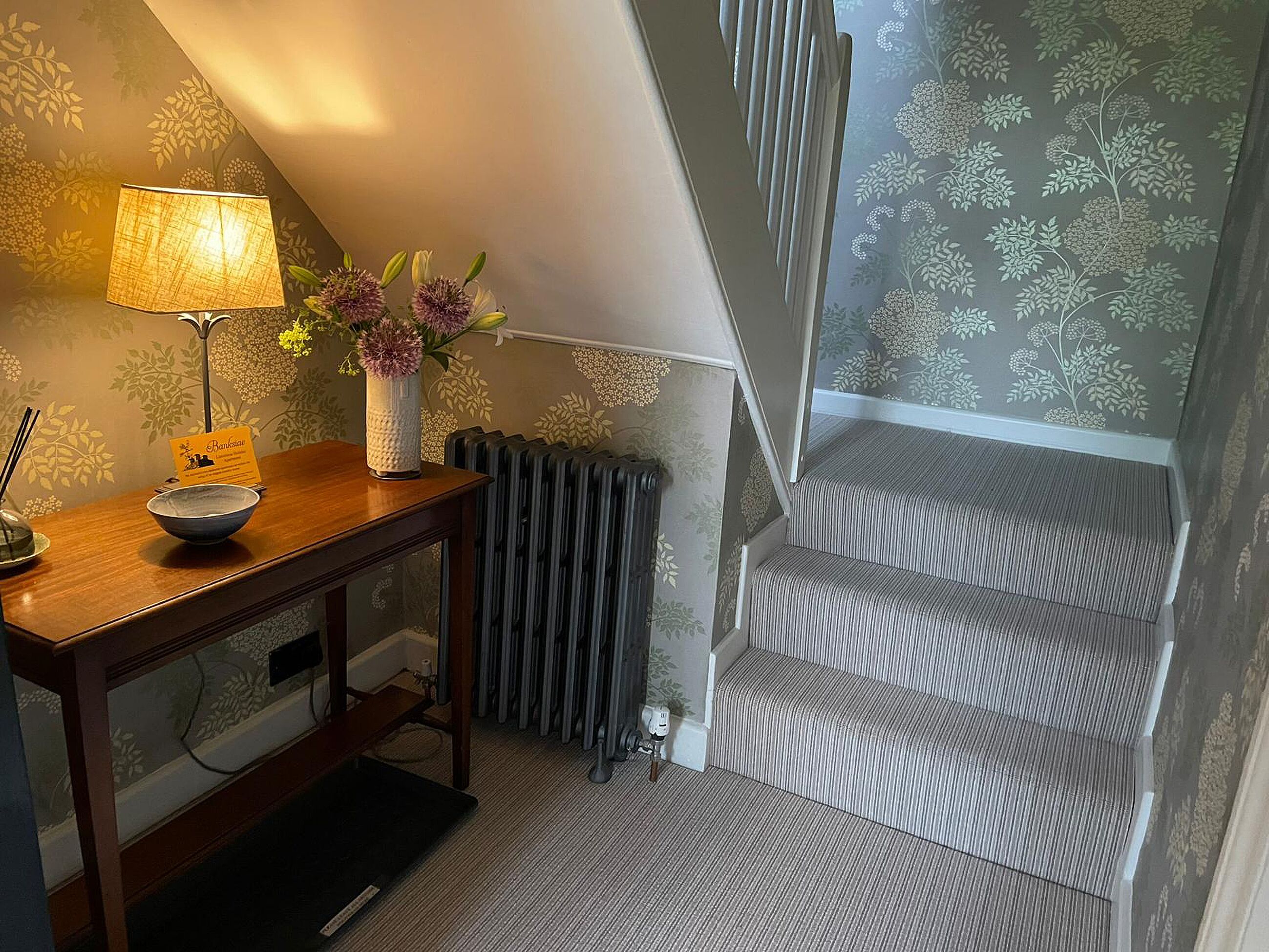 A room with a wooden desk, a lamp, a vase with flowers, a radiator, and a staircase with patterned carpet.