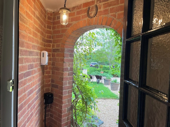 A brick-walled hallway with a hanging light fixture, a door with a glass panel, and a view of a garden with a car and greenery outside.