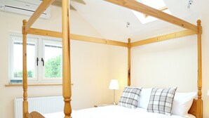 Een hemelbed met houten frame, een witte spreidek en geruite kussens. Er is een nachtkastje met lamp, een raam met zonwering en een radiator.