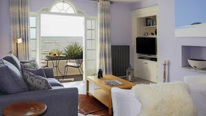 A living room with a sofa, a coffee table, a TV, and a view of the sea through an open door. - 1 bedroom accommodation in Sandgate (Sandgate)