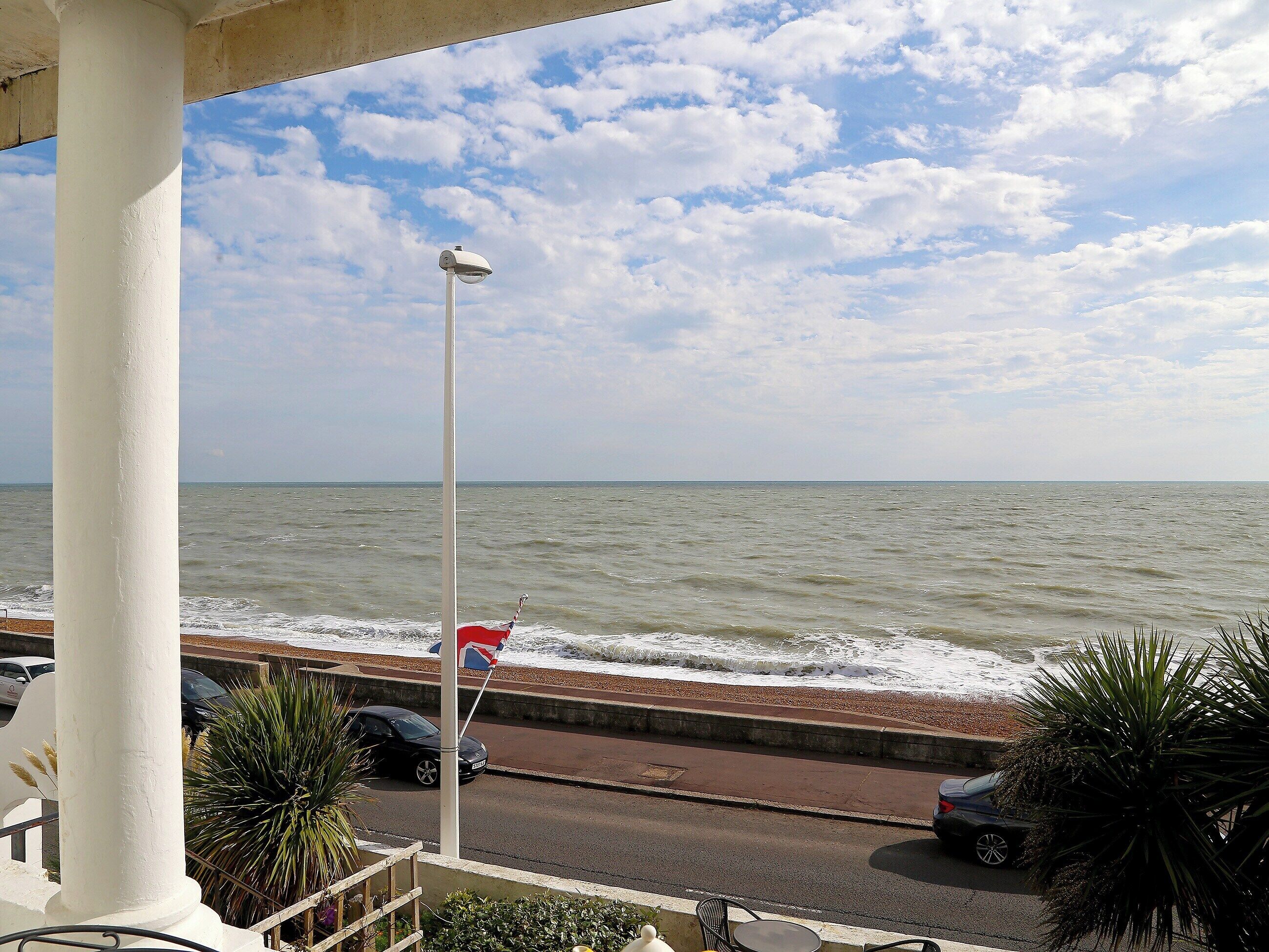 A view of the sea from a balcony with a street and parked cars in the foreground.