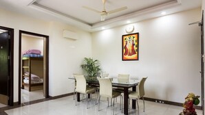 A dining area with a table and chairs, a ceiling fan, a clock, and a framed artwork on the wall. - ACHLA PARADISE – LUXURY 2BHK APARTMENT (4 BEDS) (New Delhi)