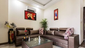 A modern living room with a brown leather sectional sofa, a glass coffee table, and two wall-mounted air conditioners. - ACHLA PARADISE – LUXURY 2BHK APARTMENT (4 BEDS) (New Delhi)