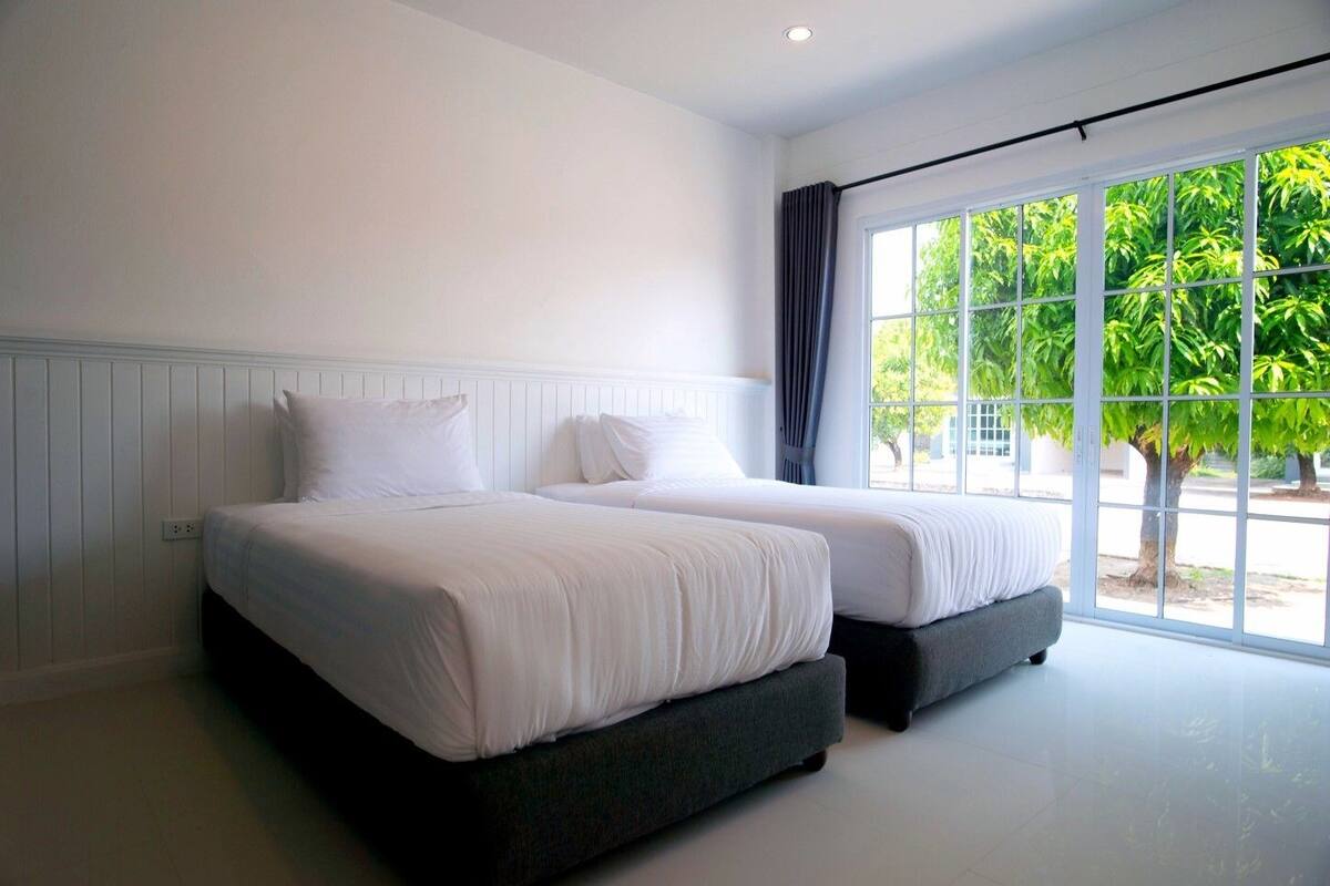 A hotel room with two beds, a large window with a view of greenery, and a white wall.