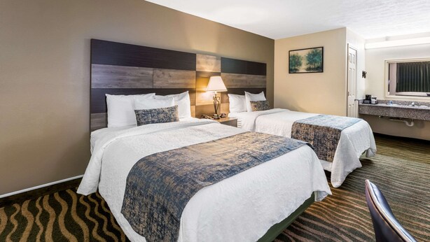 Habitación de hotel con dos camas, una mesita de noche con lámpara, un cuadro en la pared y un baño con espejo y lavamanos.