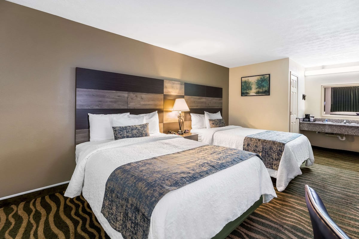 Habitación de hotel con dos camas, una mesita de noche con lámpara, un cuadro en la pared y un baño con espejo y lavamanos.