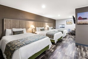 A hotel room with three beds, each with a white bedspread and a patterned throw. The room has a wooden headboard, a flat-screen TV, and a desk with a chair.