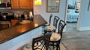 A kitchen with wooden cabinets, stainless steel appliances, and a granite countertop. There are two bar stools with patterned cushions. A framed picture hangs on the wall above the counter.