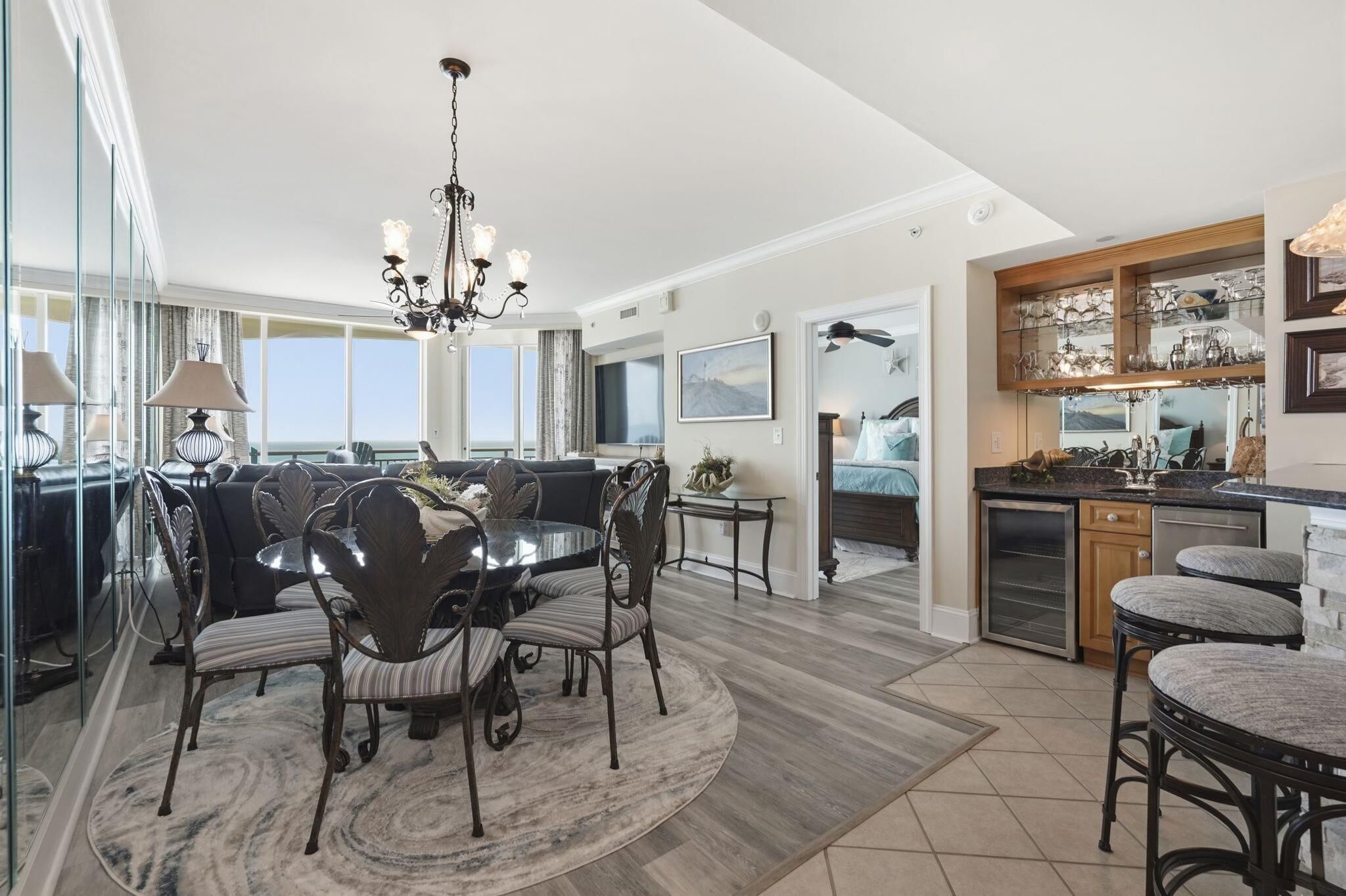 A spacious living area with a dining table, chairs, and a chandelier. There is a kitchen with a countertop and bar stools.