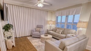 A living room with a beige sofa, a grey armchair, a round coffee table, and a large window with blinds. - MVG 1116 4 Bedroom Ocean View (North Myrtle Beach)