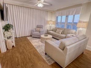 Ein Wohnzimmer mit einem beigefarbenen Sofa, einem grauen Sessel, einem runden Couchtisch und einem großen Fenster mit Jalousien.