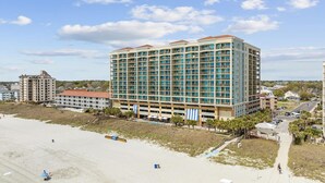 A large hotel with multiple floors and balconies, situated near a beach with a parking area and a pedestrian path. - MVG 1116 4 Bedroom Ocean View (North Myrtle Beach)