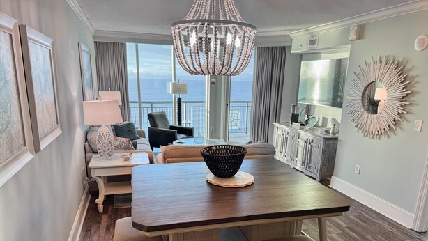 A dining area with a wooden table, cushioned stools, a chandelier, and a view of the ocean. - MVG 1116 4 Bedroom Ocean View (North Myrtle Beach)