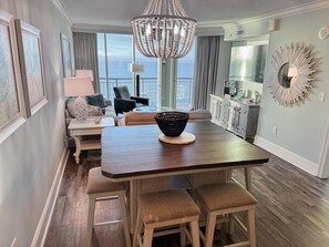 A dining area with a wooden table, cushioned stools, a chandelier, and a view of the ocean.