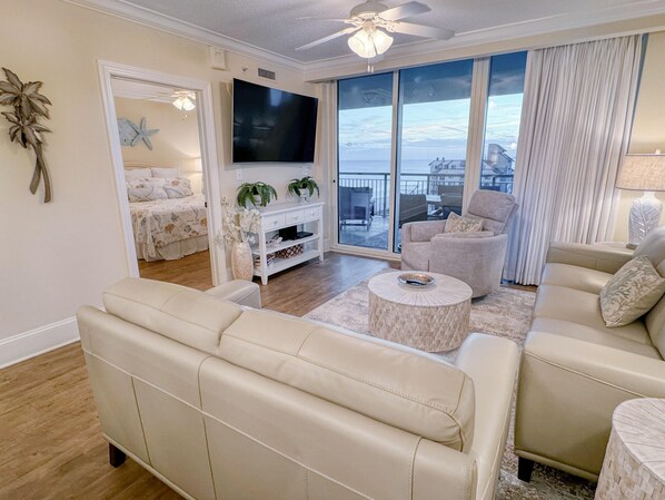 A spacious living room with a large TV, comfortable seating, a balcony with a view, and a well-lit space.