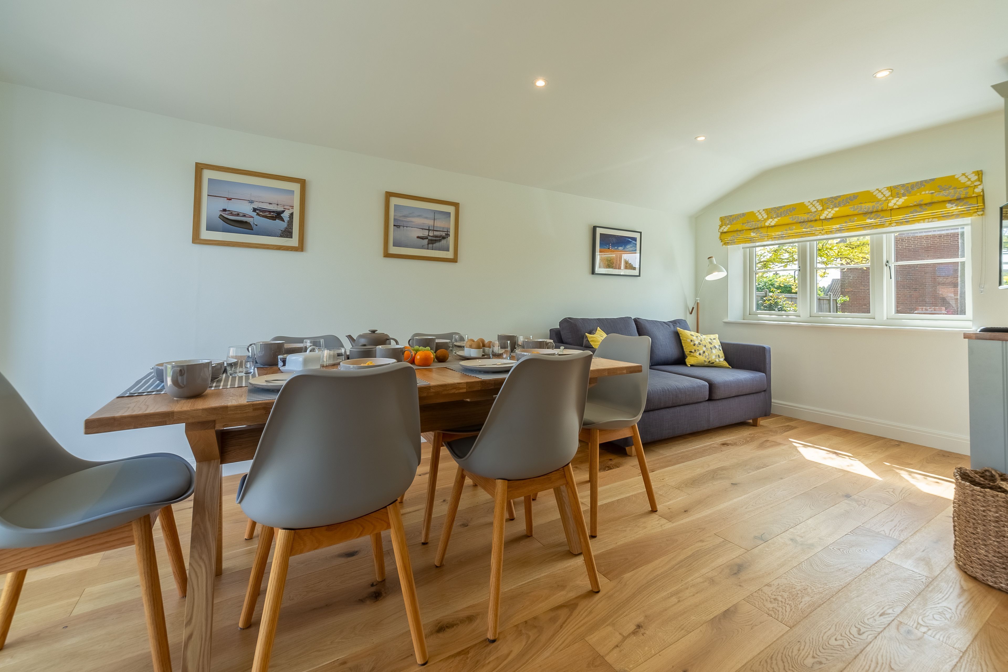 A dining area with a wooden table and grey chairs, a sofa with cushions, and a window with yellow blinds.