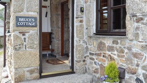Une petite maison en pierre avec une porte en bois, une enseigne indiquant « La maison de Bruce », et une petite plante en pot placée à l’extérieur.