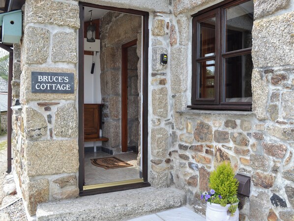 A stone cottage with a wooden door, a sign reading "Bruce's Cottage," and a small potted plant outside.