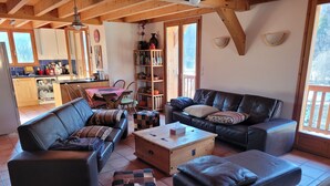 Een gezellige woonkamer met een bank, een salontafel en een beklede zitting. Een boekenplank met diverse objecten en een raam zorgen voor natuurlijk licht.