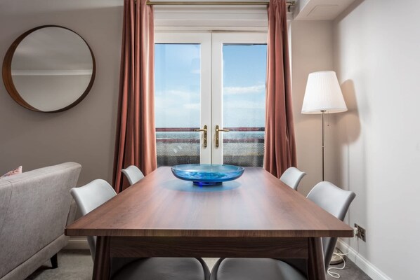 A hotel room with a wooden dining table, a blue glass bowl centerpiece, white chairs, a round mirror, and a floor lamp.