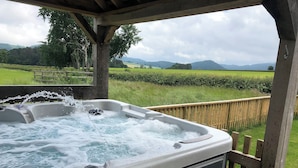 A hot tub with water jets, surrounded by a wooden deck and a view of a green landscape with hills. - Upper Close - UK10873 (Prestrigne)