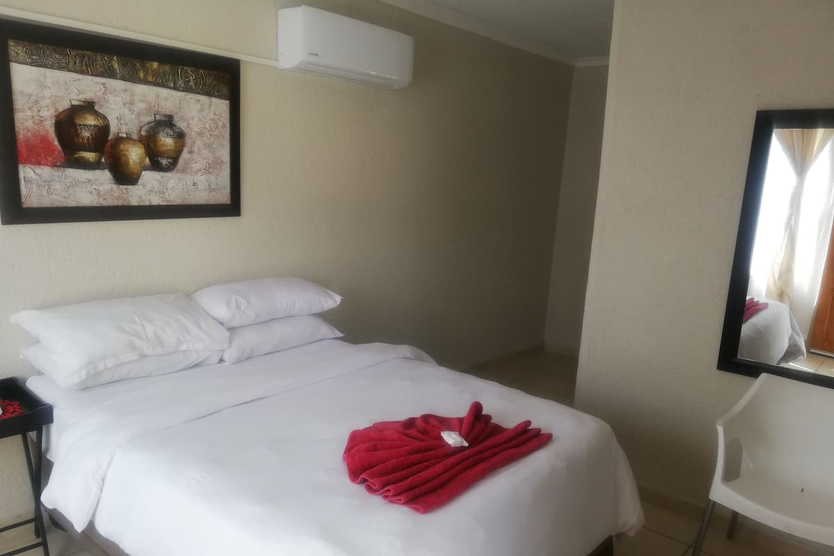 A hotel room with a bed, white pillows, a red towel, a painting, an air conditioner, and a mirror.
