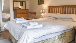 Ein ordentlich bezogenes Bett mit weißen Leinen und Handtüchern, ein hölzerner Kopfteil, ein Nachttisch mit Lampe, ein Holzsessel, eine Kommode und ein Spiegel.