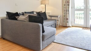 Ein graues Sofa mit mehreren Kissen, eine Stehlampe, ein kleiner Tisch und Blick durch eine Glastür.