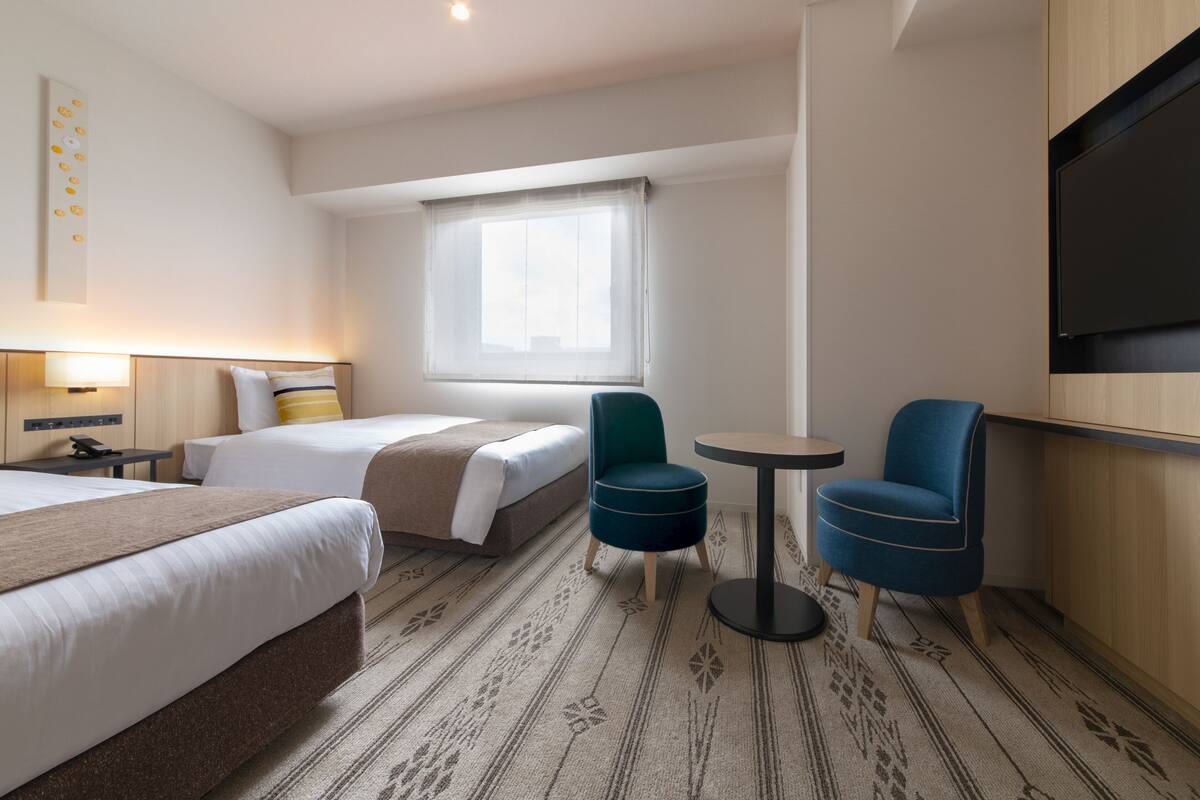Une chambre d’hôtel avec deux lits, une petite table, deux chaises, un téléviseur et une fenêtre avec un rideau léger.