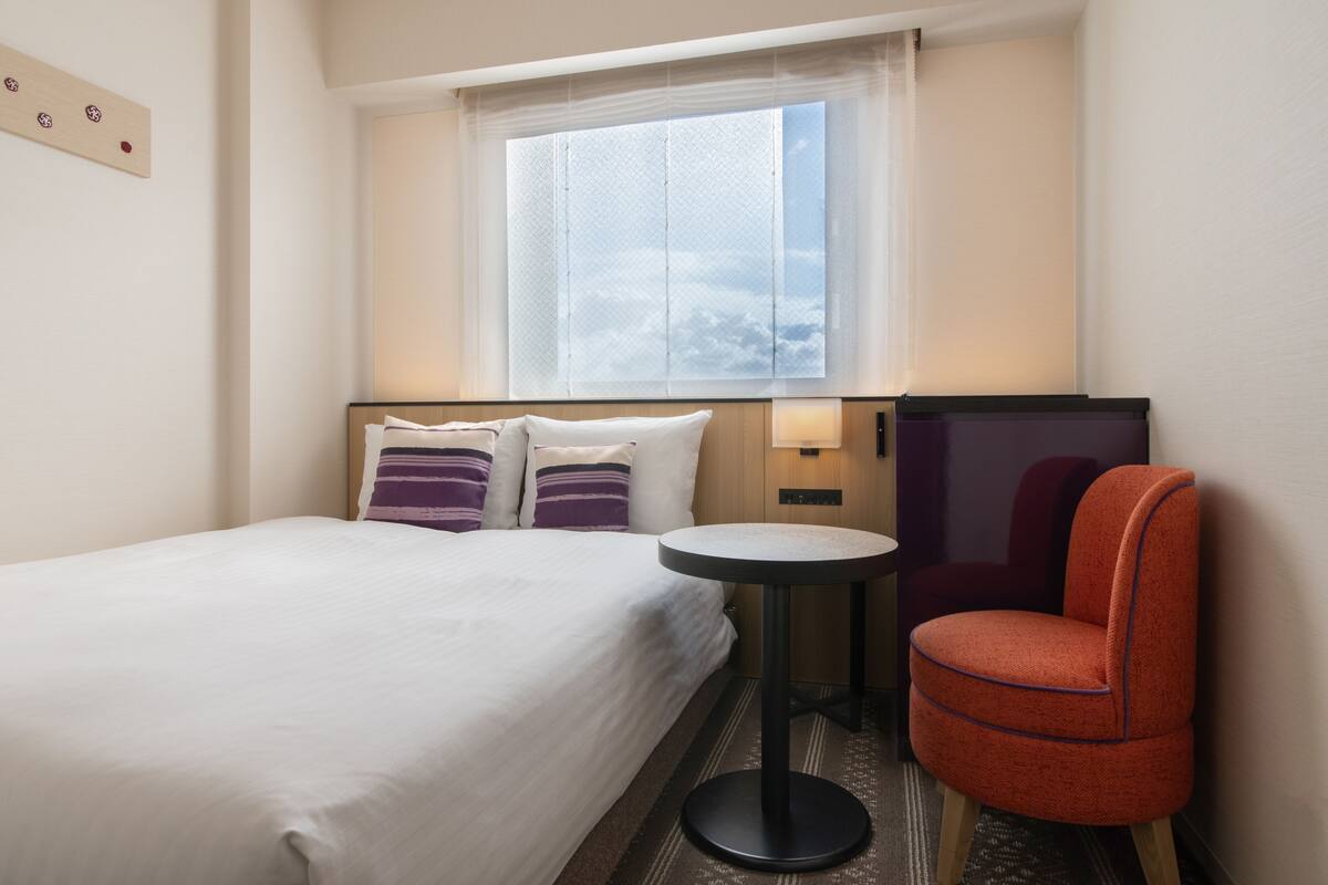 Une chambre d’hôtel avec un lit, une petite table ronde, une chaise et une fenêtre donnant sur le ciel.