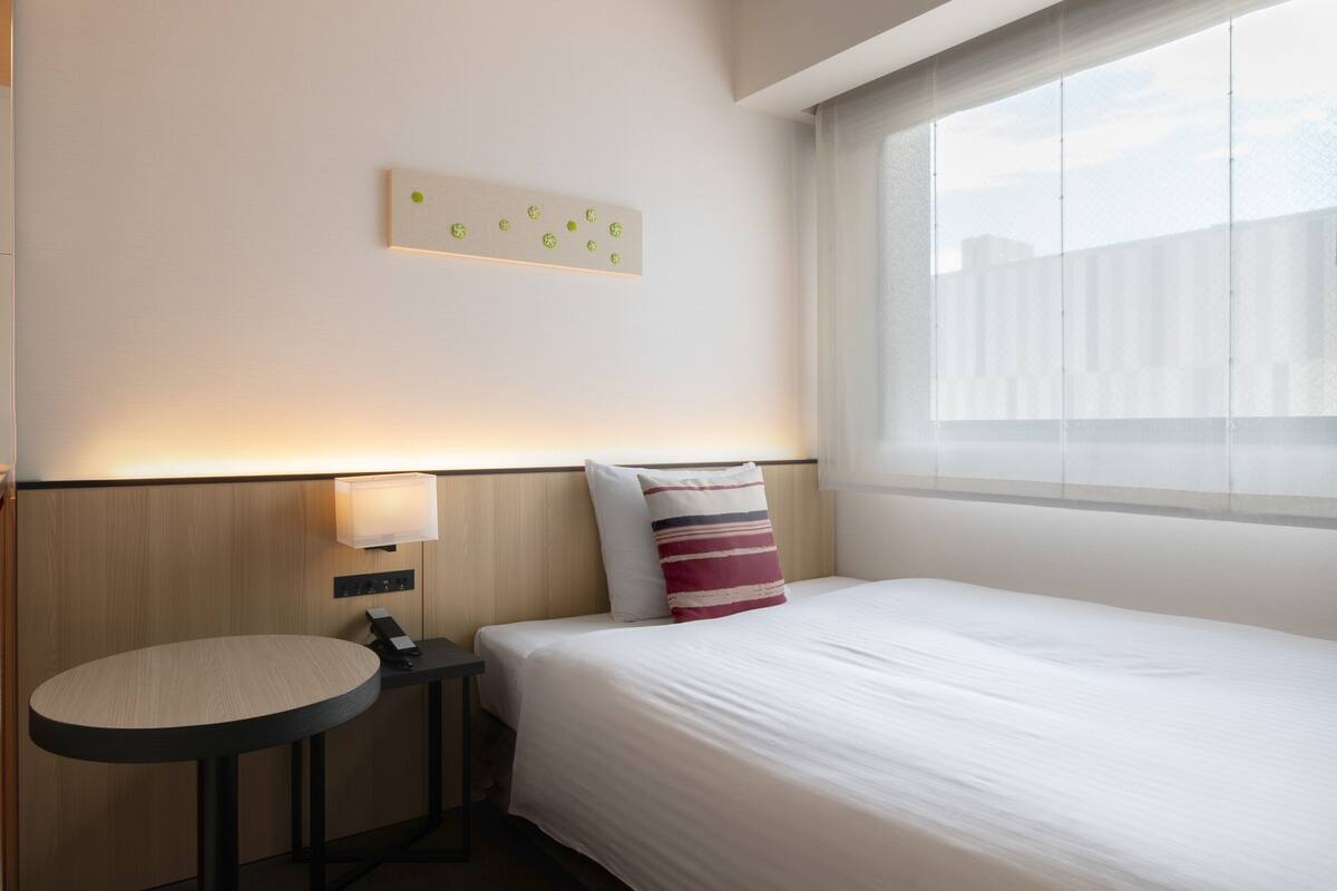 Une chambre d’hôtel avec un lit, une table de chevet avec une lampe, un luminaire fixé au mur et une fenêtre donnant sur un paysage.