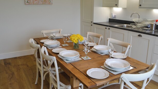 Un comedor con mesa de madera dispuesta para seis personas, sillas blancas, un florero con flores amarillas y una cocina al fondo.