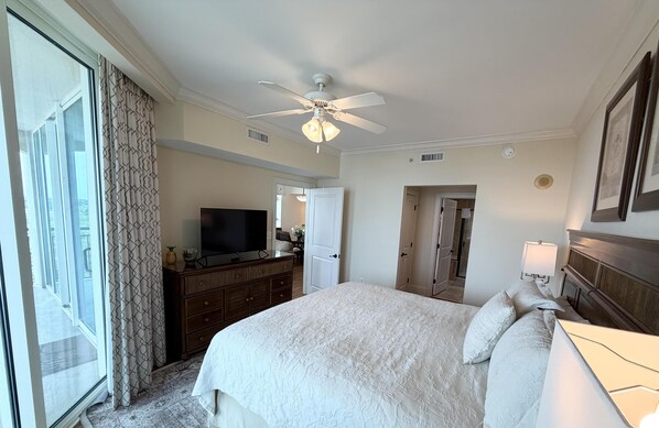 A hotel room with a bed, a ceiling fan, a television, a dresser, and a door leading to another room.