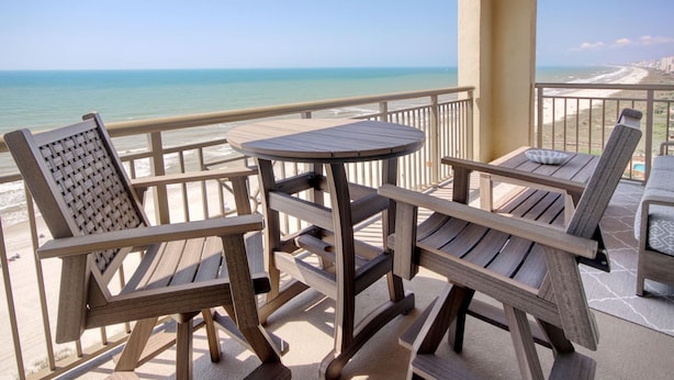 Eine Holzterrasse mit Tisch und zwei Stühlen, die auf einen Strand mit freiem Blick auf das Meer und die Küste blickt.