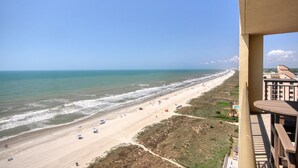 Blick auf einen Strand vom Balkon aus, mit blauem Himmel, Sandstrand und einigen Sonnenschirmen.