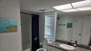 Une salle de bain avec des toilettes, un lavabo et une douche. Il y a un tableau au mur et une fenêtre avec des stores.