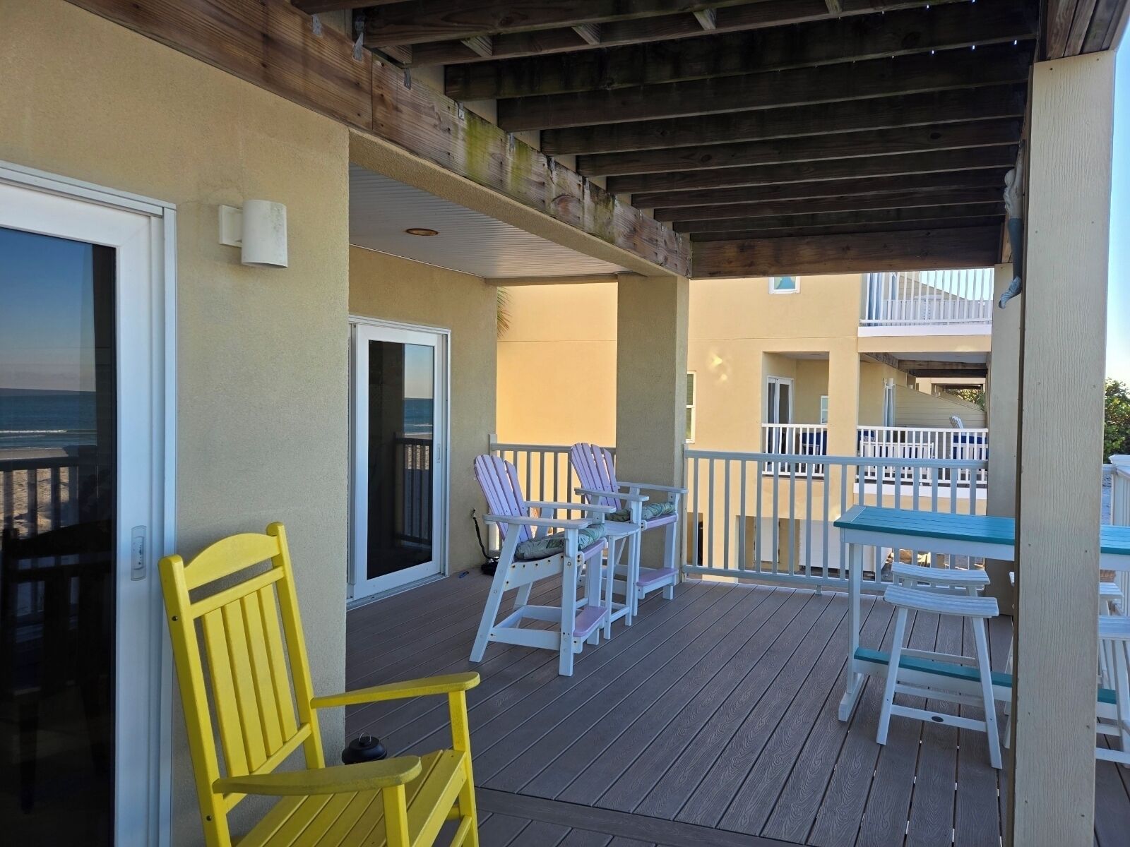 A covered outdoor deck with wooden furniture, including chairs and a table, and a view of the ocean through a glass door.