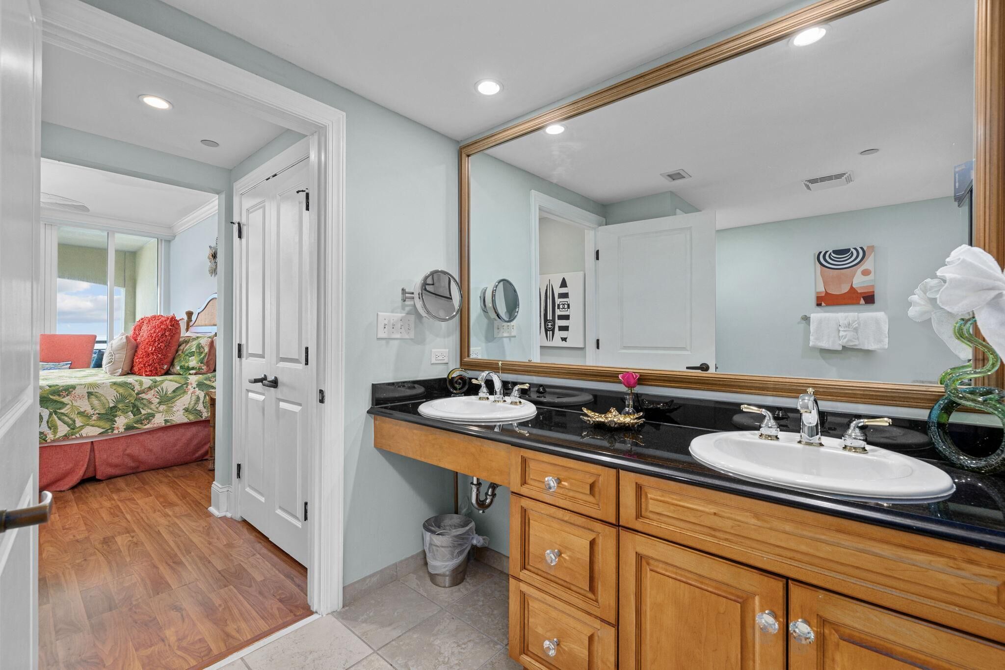 A bathroom with a large mirror, two sinks, wooden cabinets, and a view of a bedroom with a bed and a window.