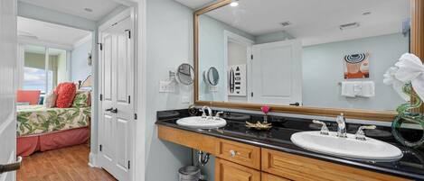 A bathroom with a large mirror, two sinks, wooden cabinets, and a view of a bedroom with a bed and a window.