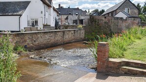 A stone wall with a small stream running alongside it, a white building with a black roof, and a brick structure with a red chimney. - 2 bedroom accommodation in Kentisbeare, Cullompton (Kentisbeare, Cullompton)